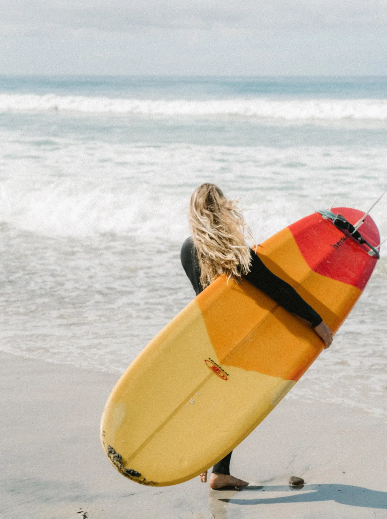 Evelyne walking into the waves with a yellow surfboard